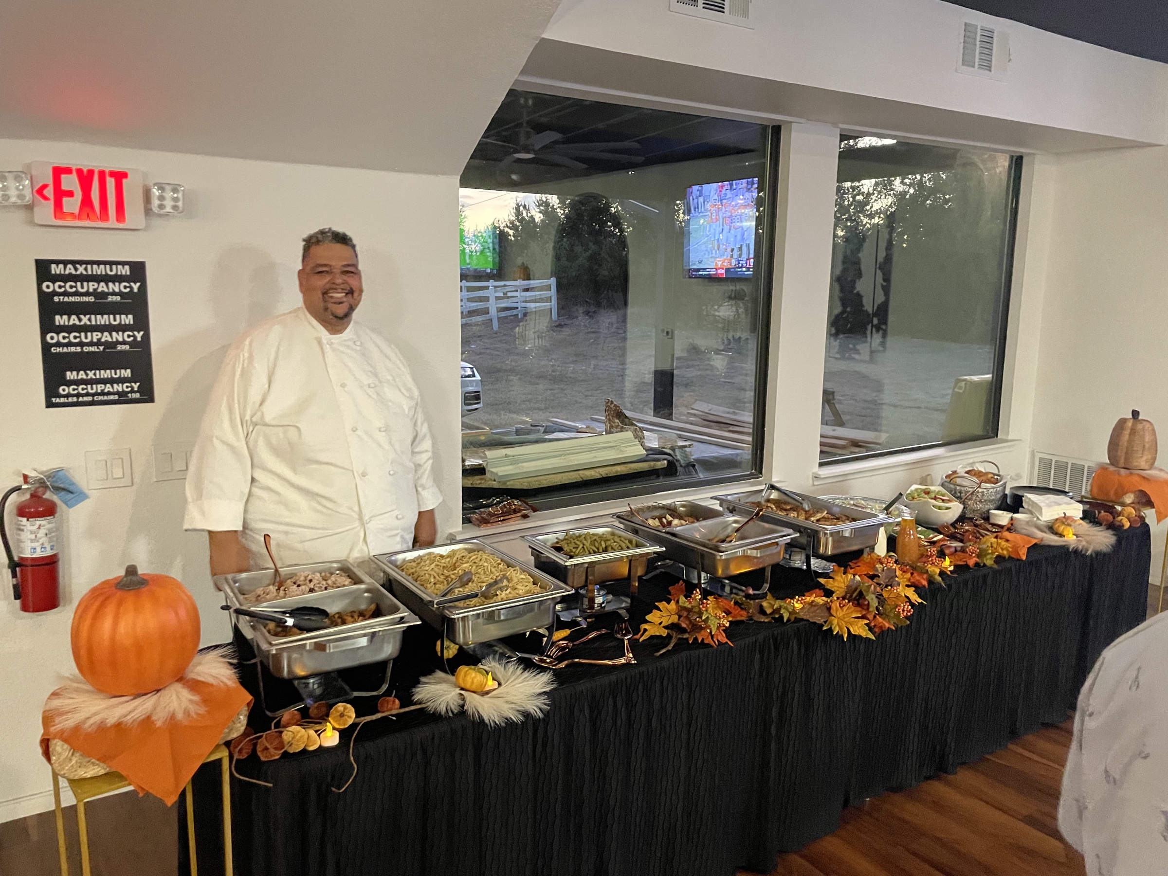 A chef in white standing beside a Thanksgiving-style holiday buffet at the venue, with chafing dishes and fall accents.